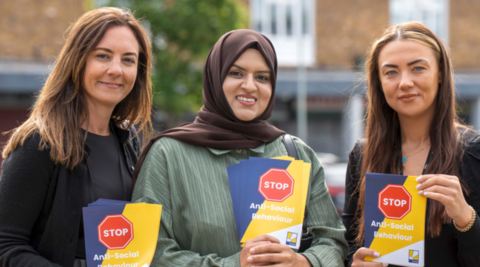 Three women are looking at the camera and holding leaflets including the words stop anti-social behaviour. A building in the background is blurred.