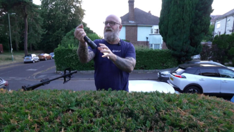 A man wearing a purple shirt holding up a ring he has just found on top of a bush.