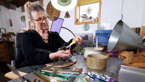 A woman with blonde hair pulled back off her face and glasses in a dark top with a large bead necklace works over a blowtorch in a studio littered with items to help her artwork
