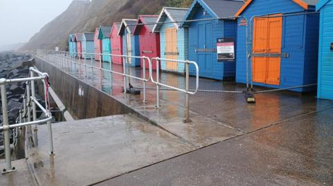 A row of multi-coloured beach huts, along a promenade, with railings along the walkway and a fence erected. Stairs lead down to the beach.