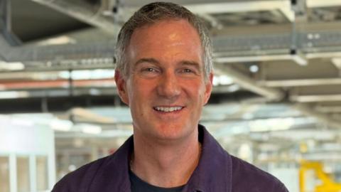 Peter Kyle stand with the Rolls-Royce manufacturing floor behind him. He is smiling and has grey hair and blue eyes. He wears a black t-shirt beneath a purple jacket. 