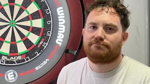 Lee Wright has curly brown hair and a beard. He is wearing a white T-shirt in front of an illuminated darts board mounted on a burgundy wall.