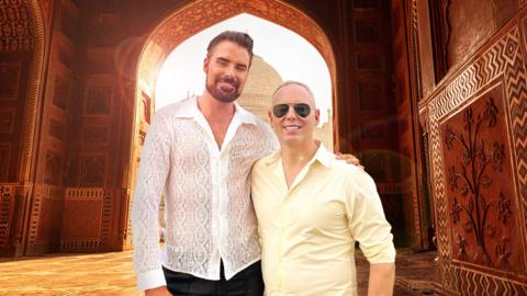 Rylan Clark and Rob Rinder standing together with sun in background of indian landmark 