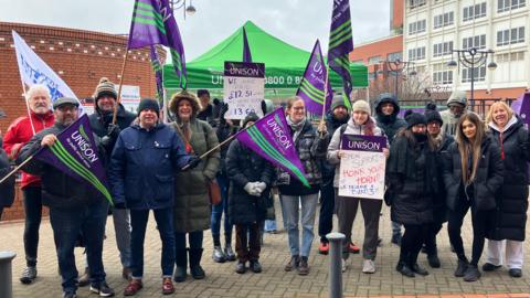 A group of union members stand outside The Leeds General Infirmary, holding UNISON flags and signs calling for fair pay.