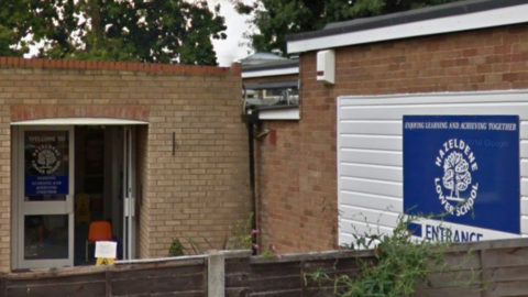 The front entrance of a school with a sign that reads "Hazeldene Lower School". There is a wooden fence in the foreground.