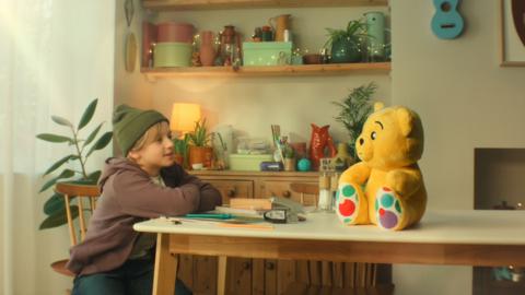 A young boy wearing a green hat and a brown hoodie is sitting at a table and looking towards Pudsey Bear who is sitting in the middle of the table. 