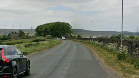 A quiet country road with a dry stone wall on the right and fields and wildflowers in the distance. On the left is a black 4x4 and a white van in the distance