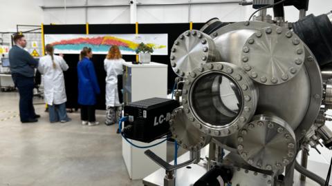 Four research scientists stand behind a shiny steel scientific device. It has five discs, the central one of which has a glass window. The scientists, two in white lab coats, stand behind it looking at a colourful periodic table stuck on a wall.