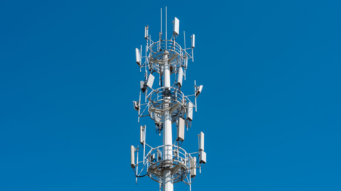 A telecommunications tower fitted with multiple antenna panels rises against a clear, bright blue sky. The structure is tall and metallic, with several circular platforms and railings positioned at different heights. Rectangular antenna units extend outward in multiple directions around the tower, along with smaller antenna rods and equipment mounted near the top. The image focuses tightly on the upper portion of the tower, with no surrounding buildings or landscape visible.