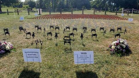 Black silhouettes representing people, cars and bicycles planted into grass. There are notices planted in front showing 1635 - the number of people killed or injured on the county's roads in 2024. There are bouquets of flowers on the left and right. There are grey dove silhouettes behind and small red county flags behind those.