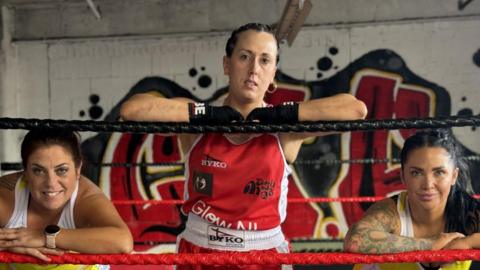 Three women standing in boxing ring with gloves on wearing sports gear. They all have dark hair pulled back behind their heads - the woman in the middle is wearing red boxing gear and is leaning on the top rope of the ring looking forward. the other two are wearing white tank tops and are leaning on the middle rope.