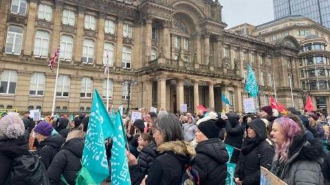 A crowd of mainly women in black coats and hats stood outside a sand coloured building, Birmingham's council house, holding blue flags.