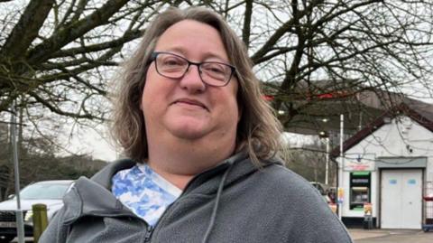 A woman with shoulder-length brown hair wearing glasses. She is wearing a blue and white floral patterned top and a grey zip-up hoodie. She is looking at the camera and smiling