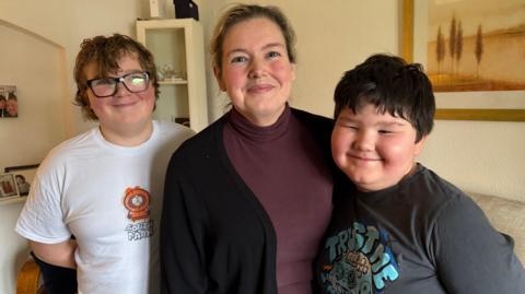 A woman is standing with her two young sons in her lounge and smiling at the camera. Behind them is a picture on the wall. The woman is wearing a black cardigan with a maroon top.