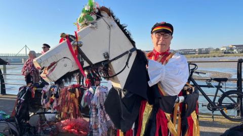 A man looks at the camera and smiles. He is wearing a Morris outfit including red and yellow silk ribbons and is holding a large wooden horse head also adorned with bells and flowers.