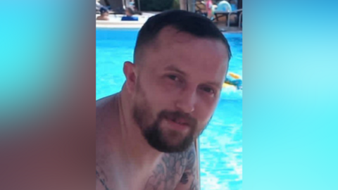 A photo of Stephen Holmes. He is shown from the shoulders up sitting in front of a pool. He has short brown hair, and a beard. He is smiling at the camera.