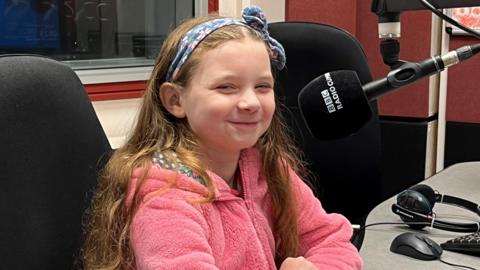 Ivy smiling and wearing a pink fluffy top and blue headband with a bow sitting in the recording studio of BBC Radio Cumbria. There is a microphone in front of her. 