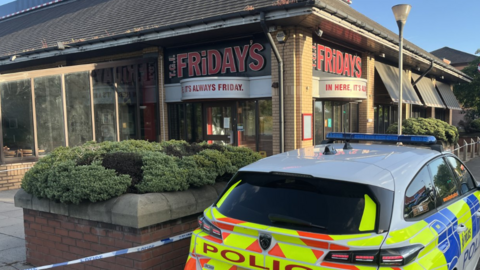 A police car outside a closed TGI Fridays restaurant. Police tape has sealed the location off.