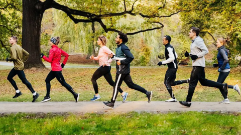 Seven people, men and women, are dressed in a variety of running gear and pictured running through a park on a tarmac path in front of trees.
