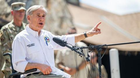 Texas Governor Greg Abbott, who uses a wheelchair, is seen seated before a microphone as he speaks and gestures. He is wearing a white shirt and khaki pants. There is a man in military fatigues slightly out of focus behind him. 