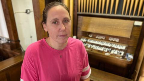 A woman in a pink t-shirt. Her hair is brown and it is tied back. She is wearing a crucifix neckless. Behind her is the damaged organ piano keys