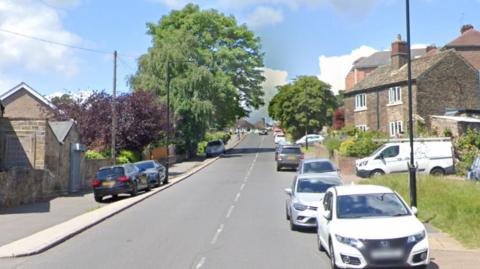 A road leading uphill. Cars parked on either side of the road. Houses on both sides of the road and trees on the side of the road.