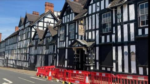 A black and white timber-framed building, there are red and white plastic barriers in front of it