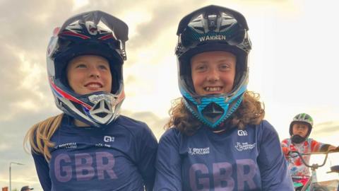 Two girls stand next to each other, while smiling and laughing. They are both wearing blue BMX gear with GBR written on it, as well as helmets. 