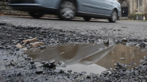 A car - slightly blurred - drives past a pothole in a main road which has a puddle in it.