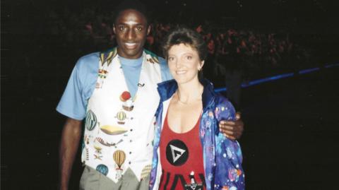 Nicola Bawden on the right and John Fashanu on the left with their arms around each other. Bawden is wearing the gladiators attire and a colourful jacket and Fashanu is wearing a blue t-shirt and a waist coat with a variety of images of hot air balloons on.