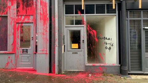 Red paint spattered across grey-coloured two doorways and the word 'borthel' and the numbers '14-16' and '4#' spraypainted in black on a glass shop window. The red paint is also on the pavement below the doors and window.  