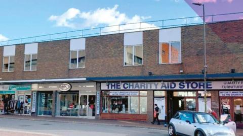 A row of shops in Camberley, including a charity shop. A mini is parked at the kerb and a pedestrian is walking past.
