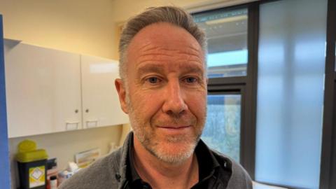 Dr Neil James, a middle-aged man with greying hair and a beard standing in his GP surgery 