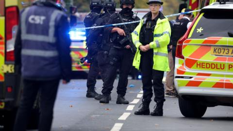 Police officers, some of whom are armed response officers, stand near police cars and other emergency vehicles behind a cordon