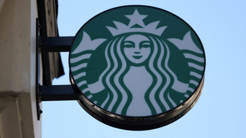 The Starbucks logo: a white mermaid on a round green background, photographed against a blue sky. 