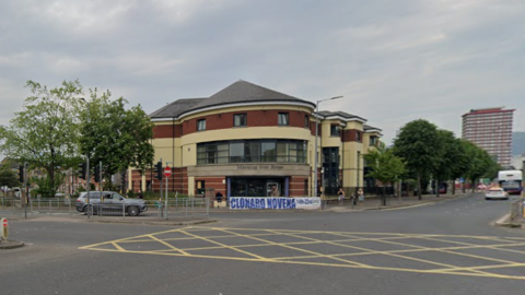 A street view of a yellow box junction. A grey jeep can be seen behind a pedestrian island with railing and traffic lights. There is a brick and yellow building in the centre behind the junction. Trees can be seen along the roadside.