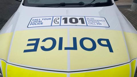 The bonnet of a police car. It is yellow and white. It has "police" written in blue block capitals backwards. It also has a large sticker on the bonnet urging people to call 101 or 999. 