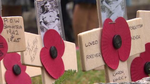 Wooden handmade crosses with pictures of men from the war, in the middle are paper poppies, all are written on with personal messages. They are sticking out of the ground.