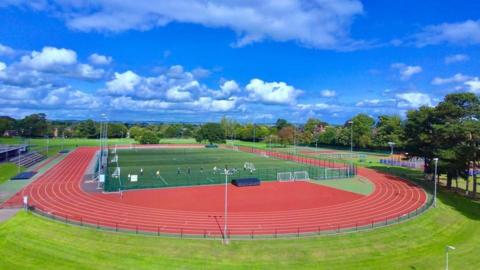 A 3G football pitch in the middle of a running track with stands to the left and tennis courts to the right. Trees can be seen in the distance under a blue sky.