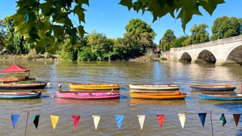 Multi-coloured boats lined up in the river with coloured bunting int the foreground, a bridge to the right and blue sky above