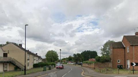 White houses on one side of the road, as well as lamp-posts and a red car and a couple of green bins. On the right hand side of the road are several brown buildings. 