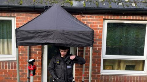 A police officer stood outside a flat in Dorking. A gazebo is placed over the door to cover it.