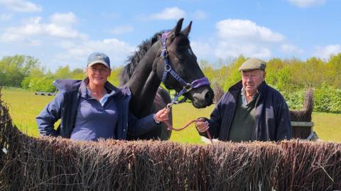 Angela Winter and "Grandad Bob" standing either side of a black horse. Angela is wearing a purple polo shirt, a purple coat unzipped and a green cap. Bob is wearing a green polo shirt, a blue coat unzipped and a green flat cap. The horse is wearing a blue and purple head collar. They are standing behind a cross country fence in a field. 