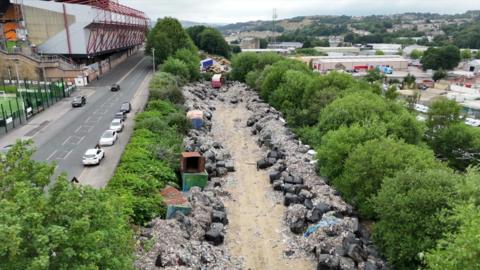 Image showing a wide track flanked by trees. Within it are hundleds of bundles of bags containing rubbish as well as rubbish which is loose. Bradford City's football stadium sits just over the road at the rear of the shot.