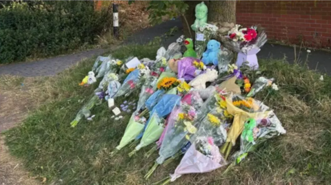 Dozens of floral tributes left next to the canal after the Jayce Olutola's death. There is a green Kermit the frog bear, and a green dinosaur teddy among the flowers as well as a blue soft bear toy.
