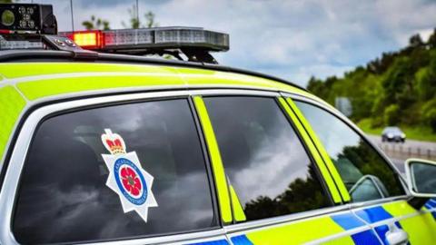 A police car is parked facing a road. It has a sticker on the back window which reads Lancashire Constabulary. A blue sky and some trees can be seen in the distance over a road.