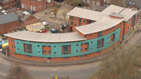 A curved apartment block with red brick walls and green cladding on the upper level. There is a curved road running alongside.