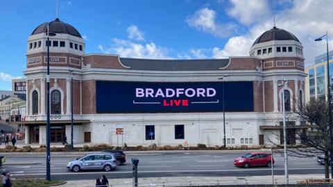 A brick and white-walled building with two domed towers and a sign reading 'Bradford Live' in white and red lettering.