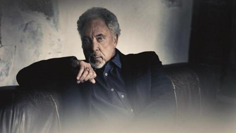 Sir Tom Jones is sitting on a leather sofa. He is wearing a dark blue jacket and shirt and his right arm is leaning on the back of the sofa.