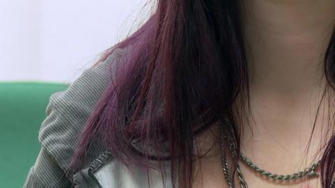 A close-up of a woman's neck. She has purple hair and is wearing a grey-hooded jacket. She is wearing a silver necklace. 